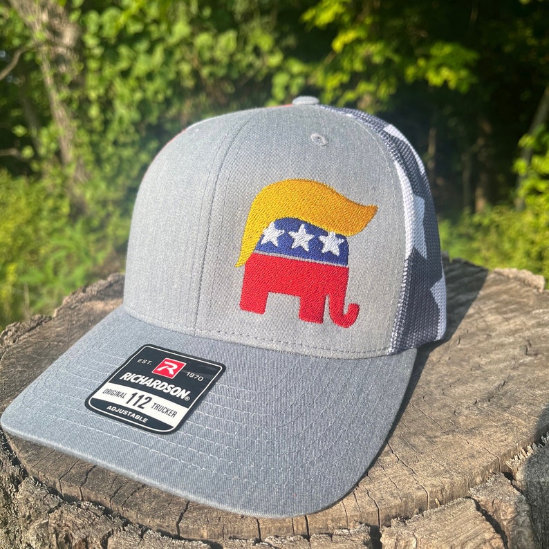 Op de afbeelding: Een grijze en zwarte trucker-pet met een rode, witte en blauwe olifant met een gele pruik. De pet heeft de tekst "EST. 1970 RICHARDSON. ORIGINAL 112 TRUCKER ADJUSTABLE" op de voorkant.