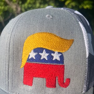 Op de afbeelding: Een grijze en witte baseballpet met een geborduurd rood, wit en blauw olifant met een gele pruik. De olifant is een symbool van de Republikeinse Partij.