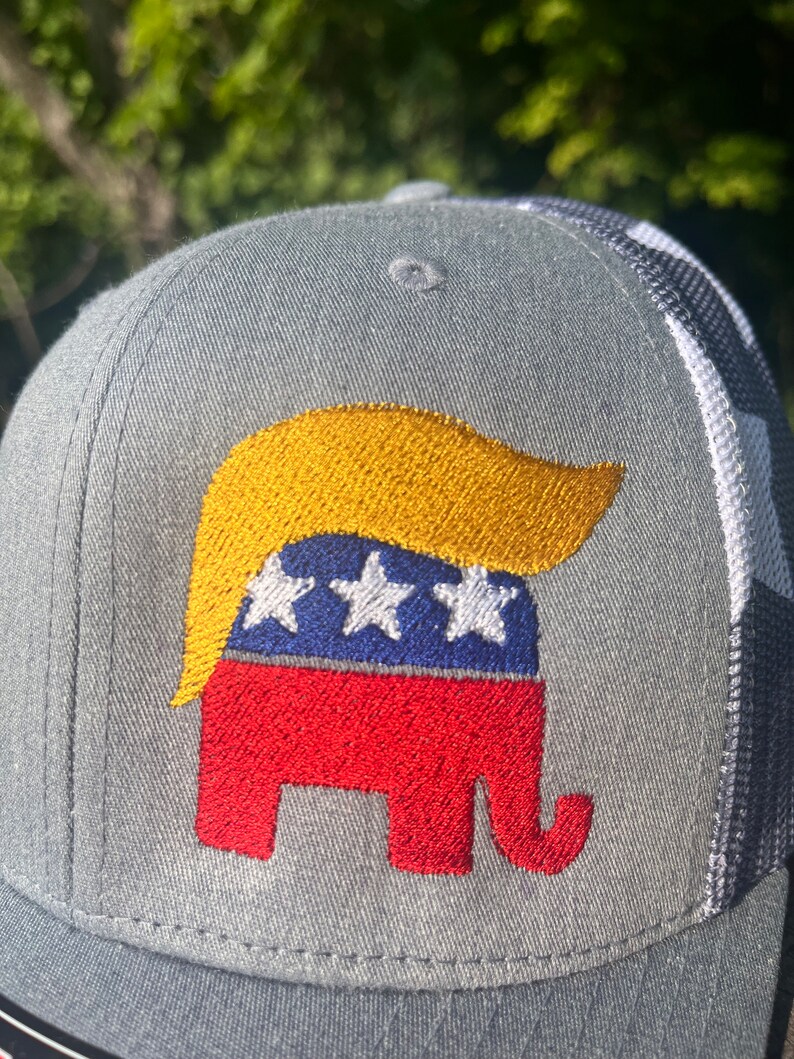 Op de afbeelding: Een grijze en witte baseballpet met een geborduurd rood, wit en blauw olifant met een gele pruik. De olifant is een symbool van de Republikeinse Partij.
