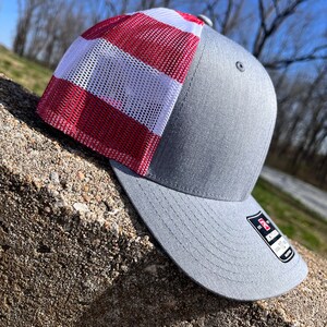 Op de afbeelding: Een grijze en rode mesh trucker-pet met een rood-wit gestreept mesh-paneel. De pet heeft een gebogen klep en een grijze stoffen kroon.