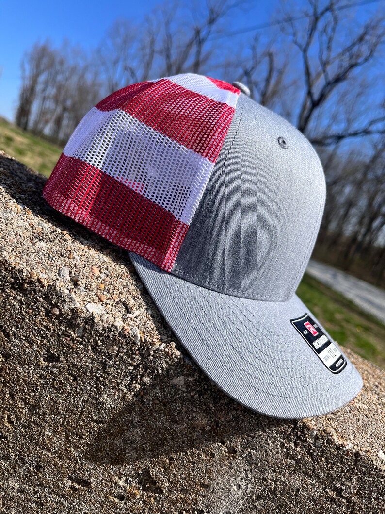Op de afbeelding: Een grijze en rode mesh trucker-pet met een rood-wit gestreept mesh-paneel. De pet heeft een gebogen klep en een grijze stoffen kroon.