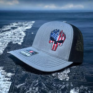 Op de afbeelding: Een grijze en zwarte truckercap met een rood, wit en blauw schedelontwerp. De schedel staat in brand en heeft een Amerikaans vlagontwerp.