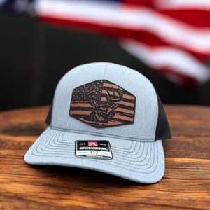May include: A grey and black baseball cap with a brown leather patch featuring a deer silhouette and an American flag design. The patch has the text "EST. 1970 RICHARDSON. 112" on it.
