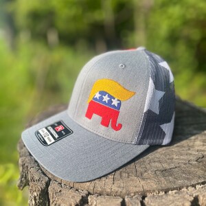 Op de afbeelding: Een grijze en witte baseballcap met een rood, wit en blauw olifantendesign. De olifant heeft een geel, golvend, haarachtig ontwerp op zijn hoofd. De pet heeft een mesh achterkant en een wit sterrenpatroon aan de achterkant.