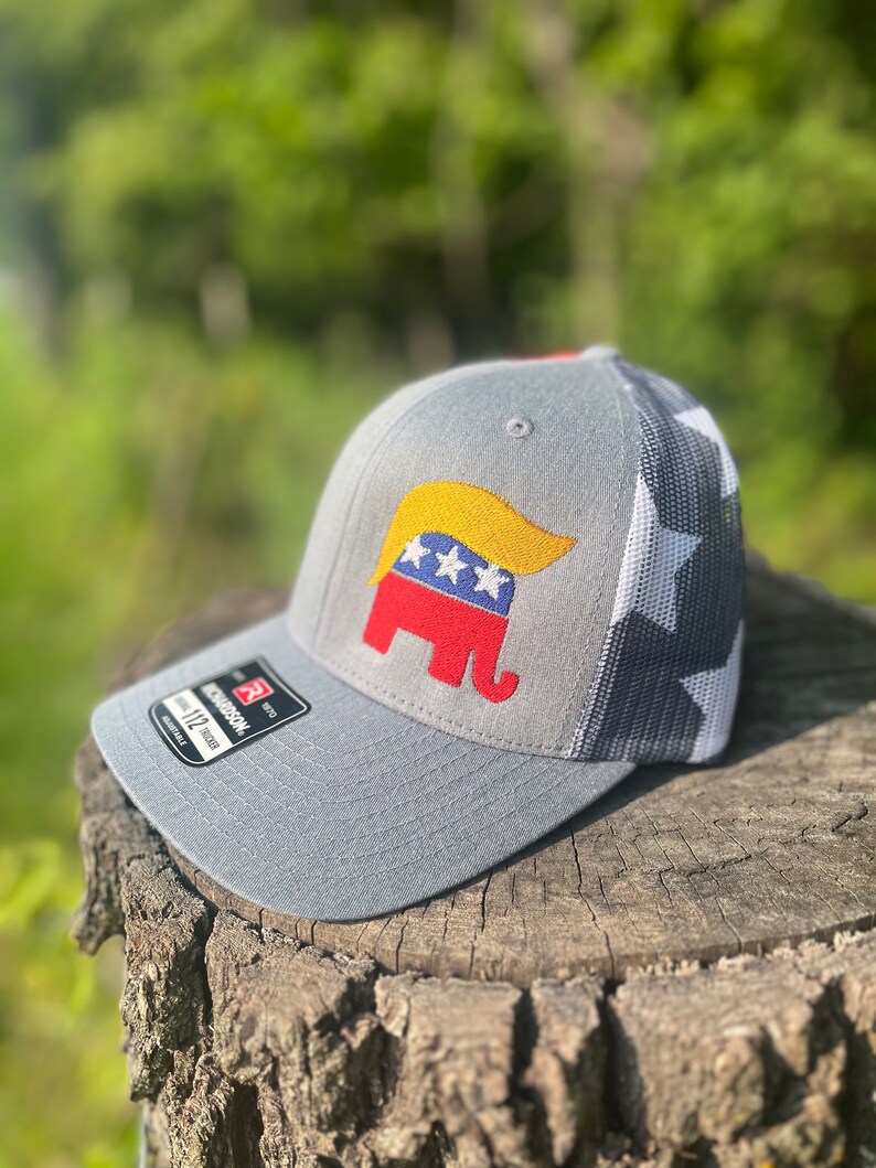 Op de afbeelding: Een grijze en witte baseballcap met een rood, wit en blauw olifantendesign. De olifant heeft een geel, golvend, haarachtig ontwerp op zijn hoofd. De pet heeft een mesh achterkant en een wit sterrenpatroon aan de achterkant.