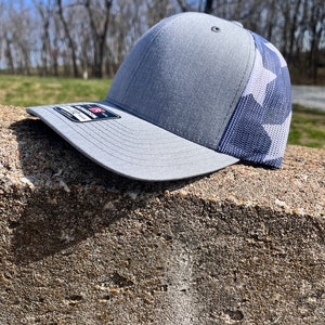 Op de afbeelding: Een grijze en witte truckercap met een mesh-rug. De pet heeft een wit sterrenpatroon op de achterkant.