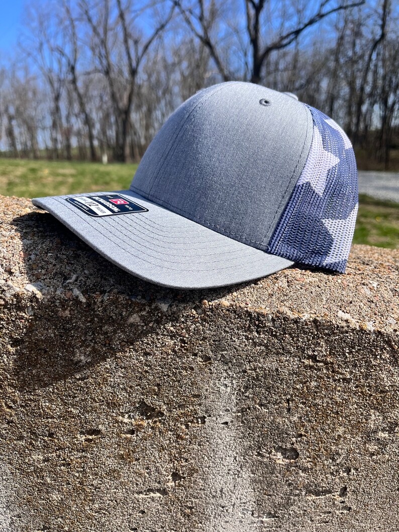 Op de afbeelding: Een grijze en witte truckercap met een mesh-rug. De pet heeft een wit sterrenpatroon op de achterkant.