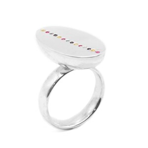 Puede incluir: Un anillo de plata con una parte superior de forma ovalada. La parte superior del anillo está decorada con una línea de pequeñas piedras preciosas de colores.