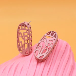 May include: Two pink oval earrings with the letters "AB ALI" cut out of the metal. The earrings are on a pink, textured surface against an orange background. The earrings are a light, dusty rose color.
