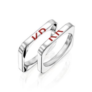 Puede incluir: Dos anillos plateados con diseño cuadrado. Cada anillo tiene las letras "KD" en esmalte rojo. Los anillos se muestran sobre un fondo blanco, resaltando sus superficies pulidas y reflectantes. Es un diseño de joyería moderno.