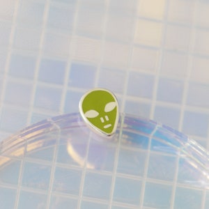 Op de afbeelding: Een groene alien-gezichtshanger met witte ogen en een mond. De hanger bevindt zich op een witte achtergrond met een rasterpatroon.