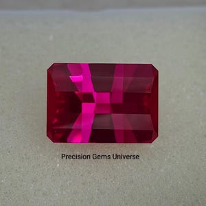 May include: A rectangular, faceted gemstone in a vibrant shade of red. The gem has a clear, polished appearance with internal reflections. The text "Precision Gems Universe" is visible below the gem.