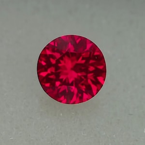 Puede incluir: Una gema de rubí redonda y facetada. La gema es de un color rojo intenso y vibrante, con múltiples facetas que reflejan la luz. La piedra preciosa está sobre un fondo neutro.