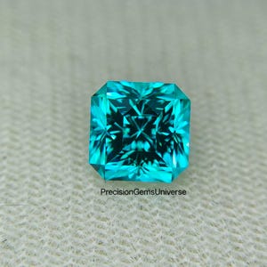 Pode incluir: Uma pedra preciosa de cor ciano vibrante com corte quadrado e facetas internas complexas que criam um padrão brilhante e reflexivo. A pedra exibe um tom azul-esverdeado saturado e está marcada com o texto PrecisionGemsUniverse.