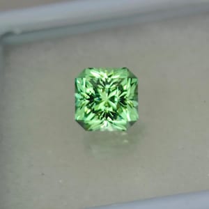 May include: A square-shaped green gemstone with a faceted cut. The gemstone has a light green color with a slight yellow hue.