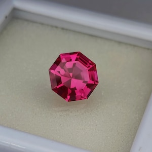 May include: A vibrant pink octagon-shaped gemstone with a faceted cut. The gemstone is displayed in a white jewelry box.
