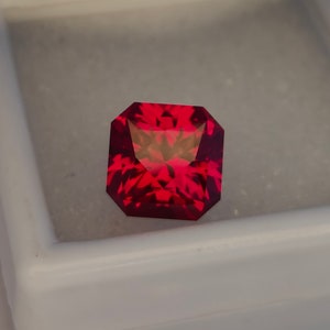May include: A deep red gemstone with a square shape and a faceted cut. The gemstone is set in a white box.