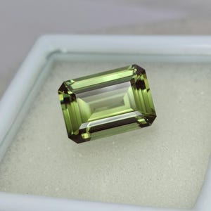 May include: A green emerald cut gemstone with a slight brown hue. The gemstone is set in a white box.
