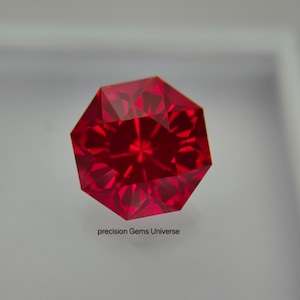Puede incluir: Una piedra preciosa de color rojo vibrante con un corte octogonal facetado, que muestra complejos reflejos de luz geométricos en toda su superficie. El texto precision Gems Universe está impreso en letras negras pequeñas debajo de la piedra.