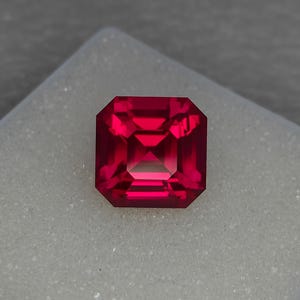 Peut inclure: Une pierre précieuse rouge foncé avec une taille émeraude carrée. La pierre précieuse est posée sur un fond blanc.