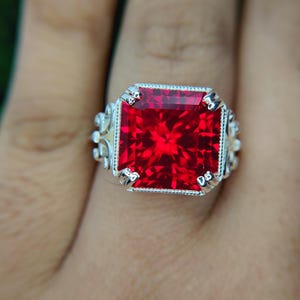 May include: A silver ring featuring a large, square-cut, vibrant red gemstone held by four prongs. The setting includes a beaded border around the stone and intricate, swirling metalwork along the sides of the band, creating a detailed design.