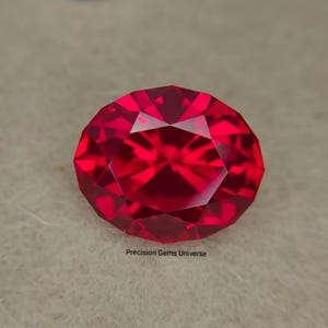 Könnte beinhalten: Ein leuchtend roter Edelstein im Ovalschliff mit einer facettierten Oberfläche, die das Licht mit hoher Klarheit reflektiert. Der Stein zeigt durchgehend einen tiefen karmesinroten Farbton. Der Text Precision Gems Universe ist unter dem Edelstein aufgedruckt.