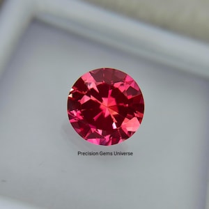 Op de afbeelding: Een ronde, gefacetteerde, levendig rode edelsteen met een briljant slijpvorm, voorzien van meerdere driehoekige en vliegervormige facetten die het licht reflecteren. De tekst Precision Gems Universe staat in het zwart onder de edelsteen.