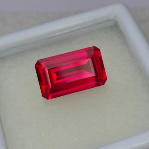 Puede incluir: Una gema roja vibrante con un corte rectangular esmeralda. La gema se muestra sobre una superficie blanca.