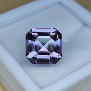 Puede incluir: Una piedra preciosa púrpura con un corte cuadrado esmeralda. La piedra preciosa está colocada en una caja blanca.