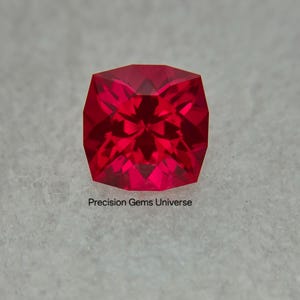May include: A vibrant, cushion-cut red gemstone with a faceted surface that reflects light. The stone is positioned above the text Precision Gems Universe, which is printed in a simple, dark, sans-serif font on a textured white surface.