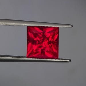 May include: A square-cut, deep red gemstone held by tweezers. The facets of the gem catch the light, creating a brilliant display. The background is a plain grey, which accentuates the gemstone's colour and form.