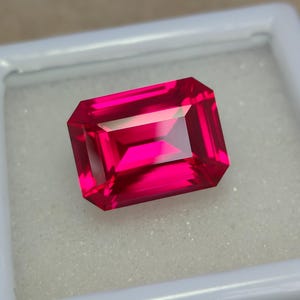 May include: A vibrant, deep red gemstone with a rectangular emerald cut. The gemstone is set in a white box.