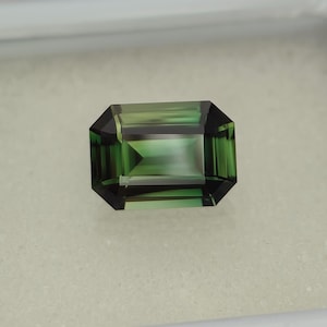 May include: Emerald-cut gemstone with a deep green hue and dark green edges. The gem has a rectangular shape with beveled corners, reflecting light and showcasing its facets. The gemstone is set against a neutral background.