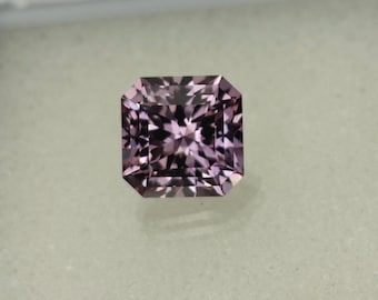 May include: A square-shaped, faceted gemstone with a purple hue. The gemstone has a star-like pattern in the center.