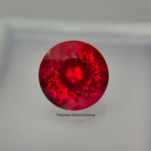 Peut inclure: Une pierre précieuse ronde à facettes d'une couleur rouge vif et profonde, présentant des reflets lumineux géométriques complexes sur toute sa surface. Le texte Precision Gems Universe est imprimé en petites lettres noires sous la pierre.