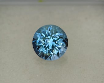 May include: A round, faceted gemstone with a brilliant cut. The gem is a vibrant shade of blue, with a complex pattern of internal reflections. The gemstone is set against a neutral background, highlighting its color and clarity.