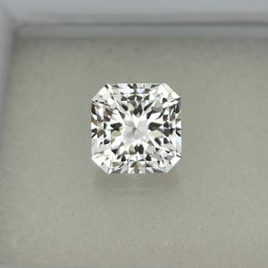 Puede incluir: Un diamante incoloro de talla radiante, de forma cuadrada con esquinas biseladas. La gema exhibe un patr&oacute;n brillante y multifac&eacute;tico, reflejando la luz. El diamante est&aacute; engastado sobre un fondo neutro, resaltando su claridad y brillo. Se trata de una joya fina.