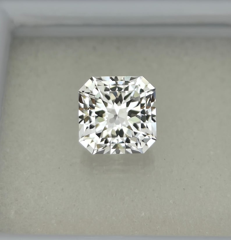 Puede incluir: Un diamante incoloro de talla radiante, de forma cuadrada con esquinas biseladas. La gema exhibe un patr&oacute;n brillante y multifac&eacute;tico, reflejando la luz. El diamante est&aacute; engastado sobre un fondo neutro, resaltando su claridad y brillo. Se trata de una joya fina.