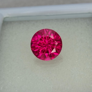 May include: A vibrant pink gemstone with a round cut and a faceted design. The gemstone is set against a white background.
