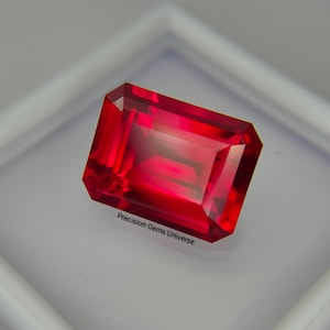 Op de afbeelding: Een levendige, rechthoekige, smaragdgeslepen rode edelsteen met scherpe facetten en hoge helderheid. De steen wordt getoond op een witte ondergrond met de tekst Precision Gems Universe in zwarte letters bij de onderste rand van de steen.