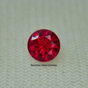 May include: A round, faceted, brilliant red gemstone. The gem is set against a neutral background. The text "Precision Gems Universe" is visible at the bottom of the image.
