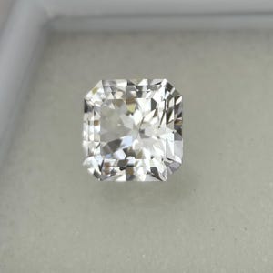 Puede incluir: Una gema incolora de corte cuadrado con una superficie facetada brillante. La gema se presenta sobre un fondo neutro, resaltando su claridad y brillo. La forma es una elecci&oacute;n cl&aacute;sica y elegante para joyer&iacute;a.