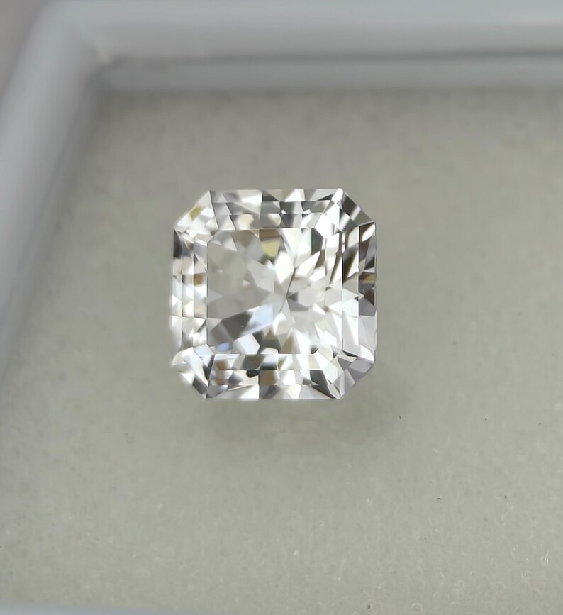 Puede incluir: Una gema incolora de corte cuadrado con una superficie facetada brillante. La gema se presenta sobre un fondo neutro, resaltando su claridad y brillo. La forma es una elecci&oacute;n cl&aacute;sica y elegante para joyer&iacute;a.
