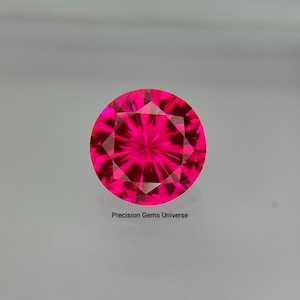 Puede incluir: Una piedra preciosa redonda facetada de un color rosa intenso vibrante con un corte brillante. La piedra presenta un patrón simétrico de facetas triangulares que irradian desde el centro. El texto Precision Gems Universe está impreso debajo.