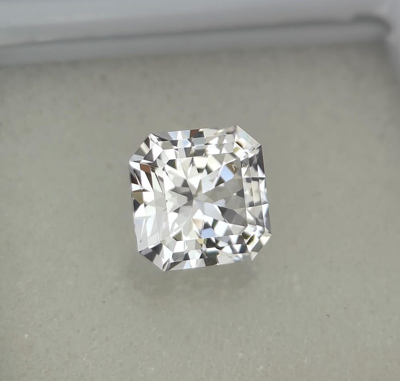 Puede incluir: Un diamante incoloro de talla radiante, de forma cuadrada y facetada. La gema se presenta sobre un fondo neutro, resaltando su brillo y claridad. Este diamante es un s&iacute;mbolo de lujo y elegancia.