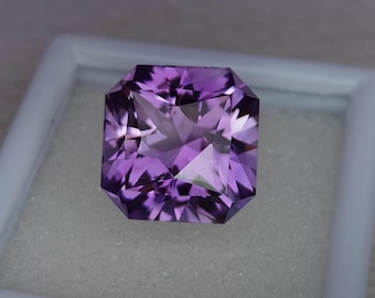May include: A square-shaped, faceted amethyst gemstone with a purple hue. The gemstone is set in a white box.