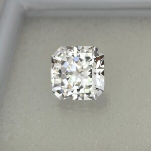 Puede incluir: Un diamante de talla radiante, de forma cuadrada y m&uacute;ltiples facetas. La gema incolora brilla con la luz, reflejando un brillo brillante. El diamante se encuentra sobre un fondo neutro, resaltando su claridad y brillo.