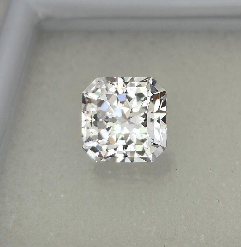 Puede incluir: Un diamante de talla radiante, de forma cuadrada y m&uacute;ltiples facetas. La gema incolora brilla con la luz, reflejando un brillo brillante. El diamante se encuentra sobre un fondo neutro, resaltando su claridad y brillo.