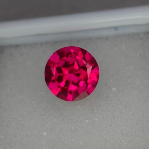 Puede incluir: Una gema roja vibrante y profunda con un corte redondo y facetado. La gema está colocada sobre un fondo blanco.