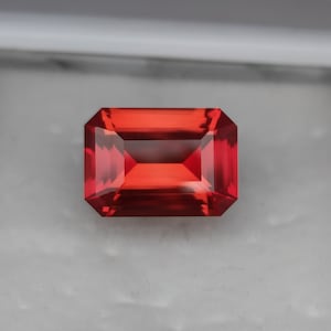 Könnte beinhalten: Ein rechteckig geschliffener, leuchtend roter Edelstein mit facettierter Oberfläche. Der Edelstein hat ein klares, poliertes Aussehen, das Licht reflektiert und seine innere Struktur zeigt. Der Edelstein ist vor einem neutralen Hintergrund platziert.